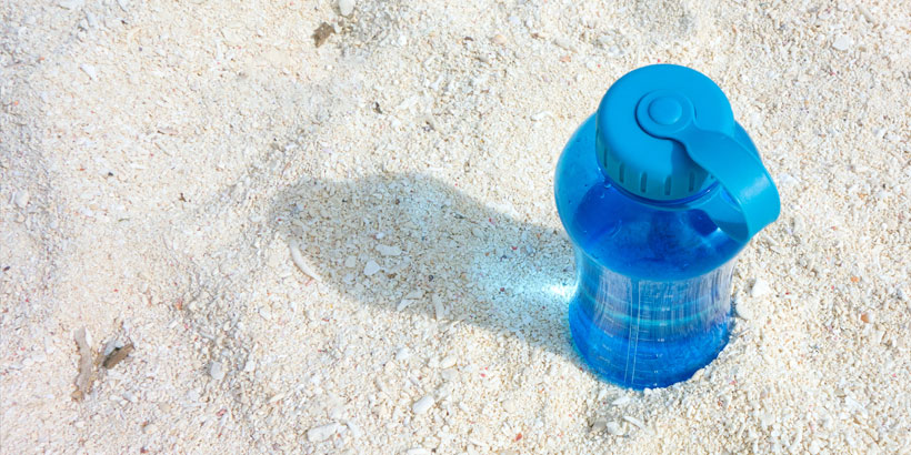 Blue reusable water bottle sitting on clean sand casting a shadow to the left.