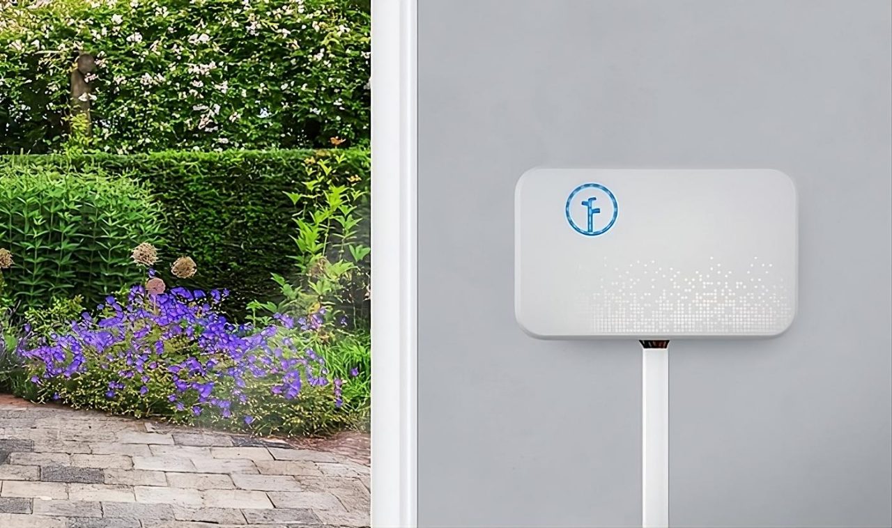 A Rachio Gen 2 sprinkler is seen on a wall next to a window with a pretty garden.